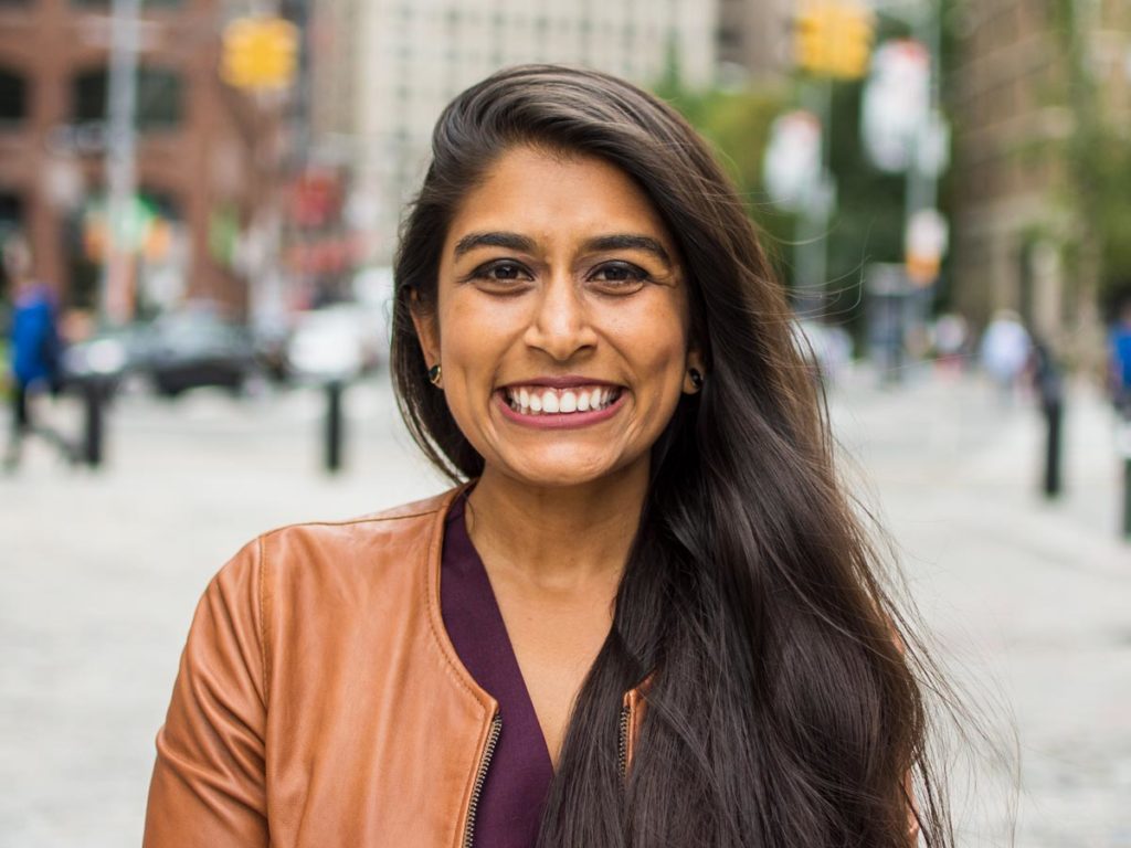 portrait of Sabrina Talukder standing outside in New York, wearing a brown coat and smiling