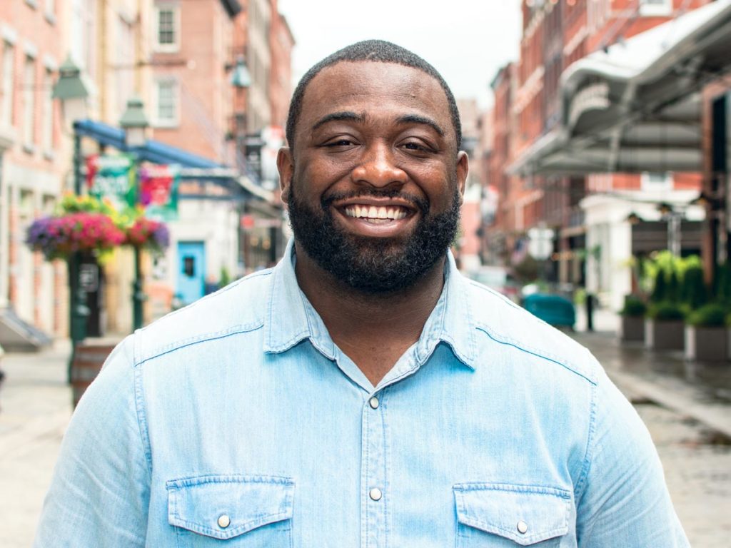 Victor Dempsey stands outside in Manhattan, smiling and wearing a blue denim shirt